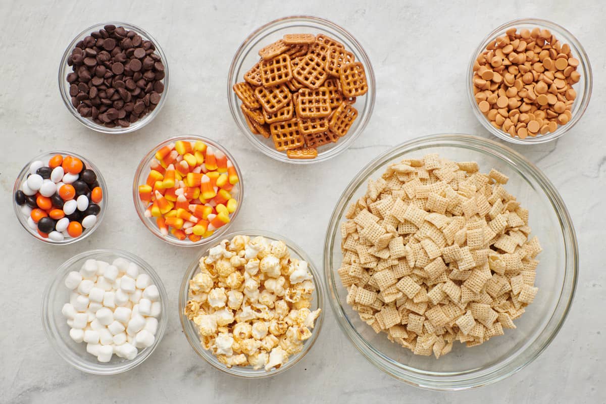 Ingredients for recipe in individual bowls: chocolate chips, candy pieces, mini marshmallows, candy corn, pretzels, popcorn, chex, and peanut butter chips.